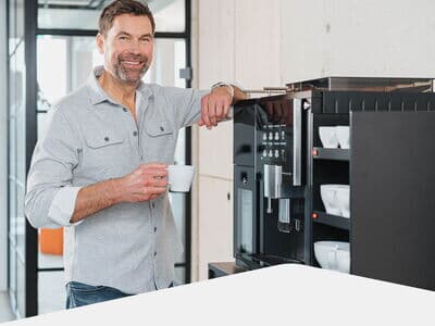 Mann mit Tasse Kaffee steht lächelnd neben dem modernem Kaffeevollautomaten im Büro