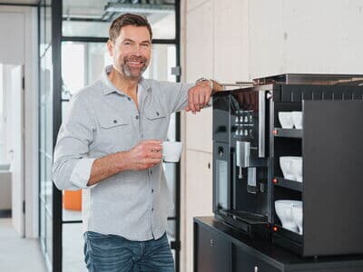 Ein Mann stützt sich in einer Büroumgebung an einem Vollautomaten ab und halt lächelnd eine Kaffeetasse