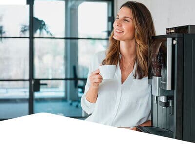 Frau lehnt am Kaffeeautomaten mit Tasse in heller Umgebung